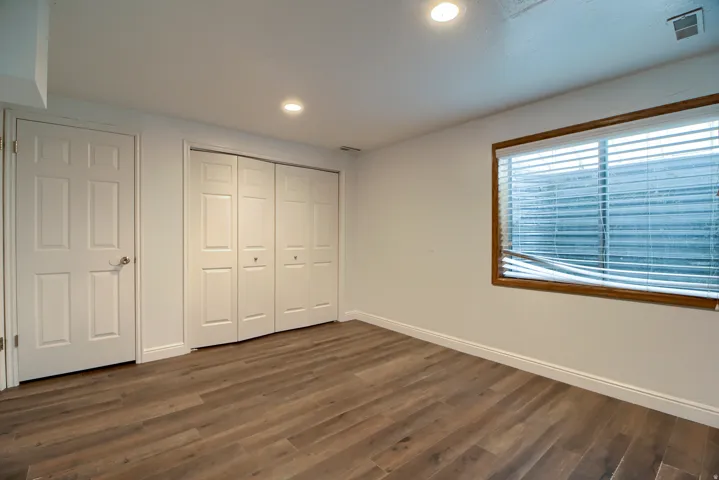 Spacious bedroom featuring warm wood-finish flooring, abundant natural light from a large picture window with blinds, and ample closet storage with dual sliding doors