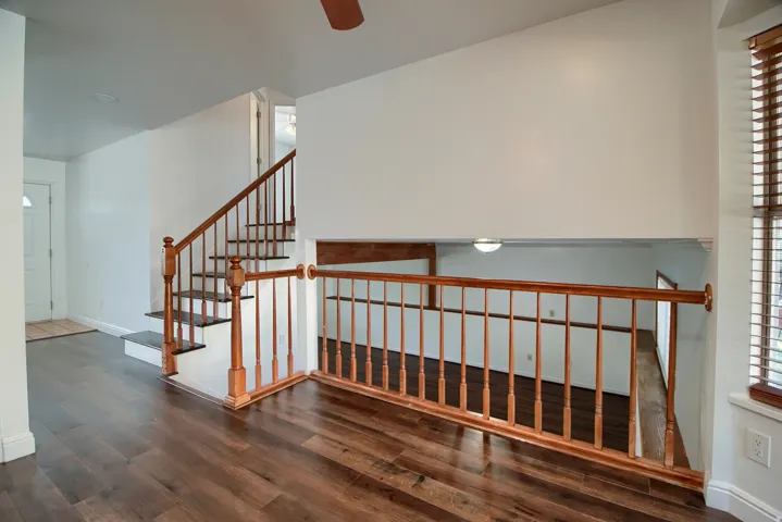 Wood-finish flooring throughout, solid wood staircase with balusters, ceiling fan fixture, white trim, and a window with wood-finish blinds