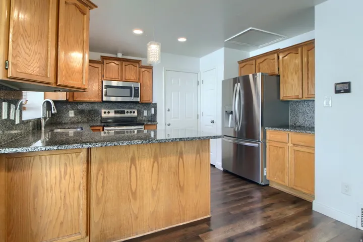 Spacious kitchen featuring warm wood cabinetry, stainless steel appliances including a French-door refrigerator, speckled stone countertops, dark wood-finish flooring, and recessed lighting throughout