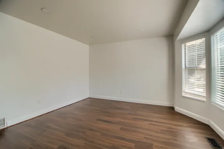 Bright corner room showcasing rich wood-finish flooring, clean white walls, and abundant natural light from dual windows with plantation shutters