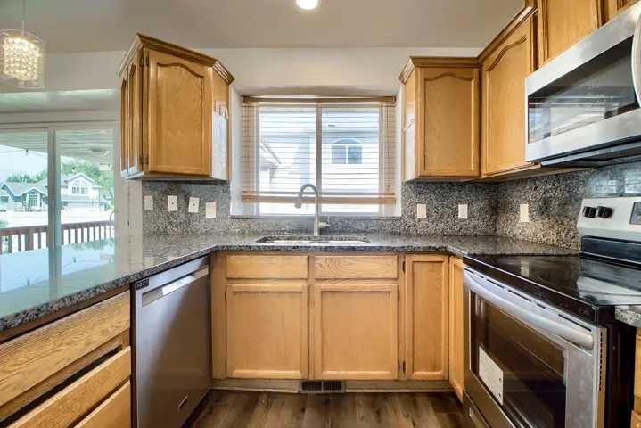 Galley kitchen featuring warm wood cabinetry with raised-panel doors, granite countertops, stainless steel appliances including double wall ovens, a center island with prep space, and a window overlooking the exterior