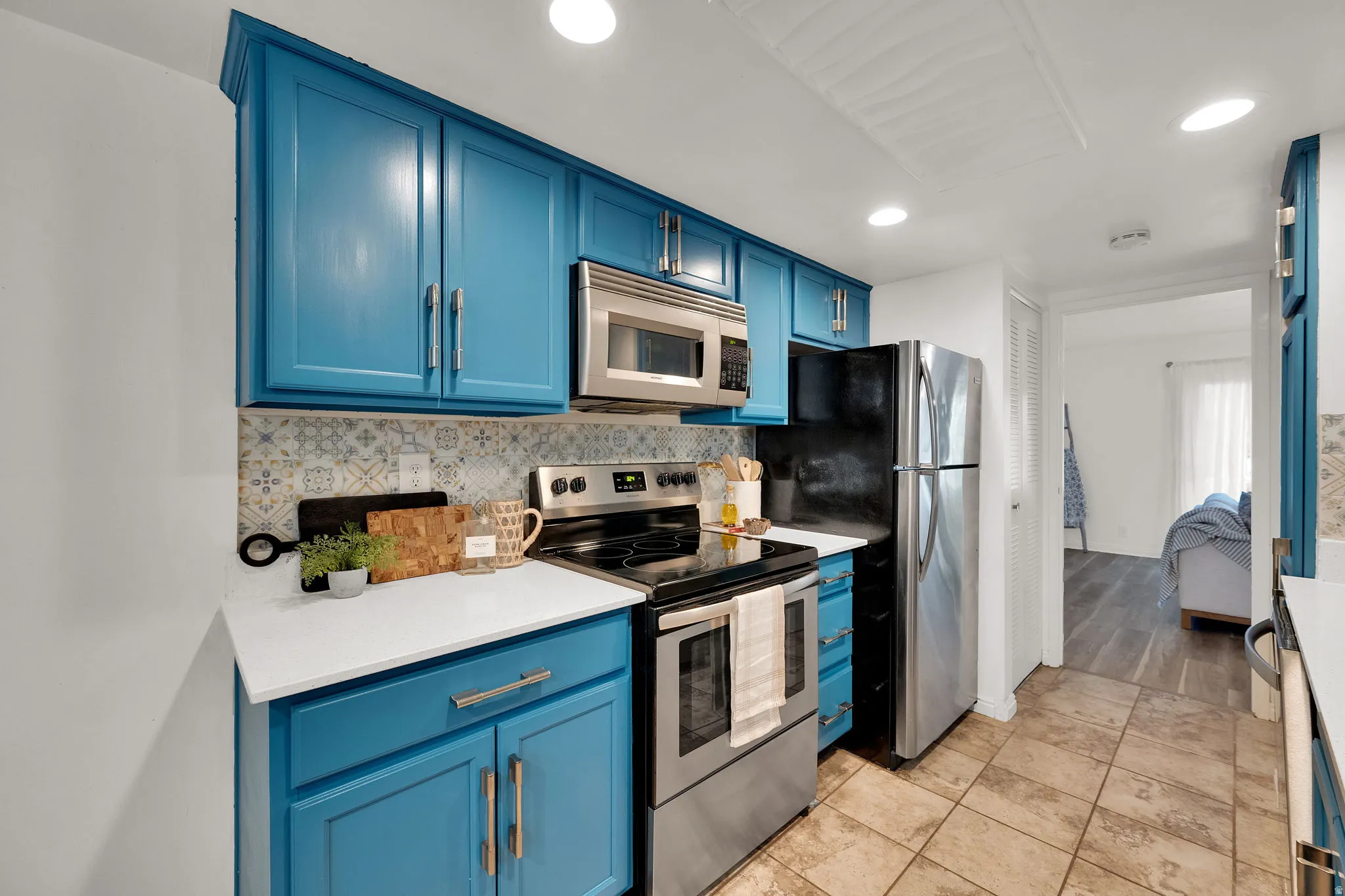 Galley kitchen featuring custom blue cabinetry with brushed metal hardware, quartz countertops, tile backsplash, and stainless steel appliances including a gas range, microwave, and refrigerator