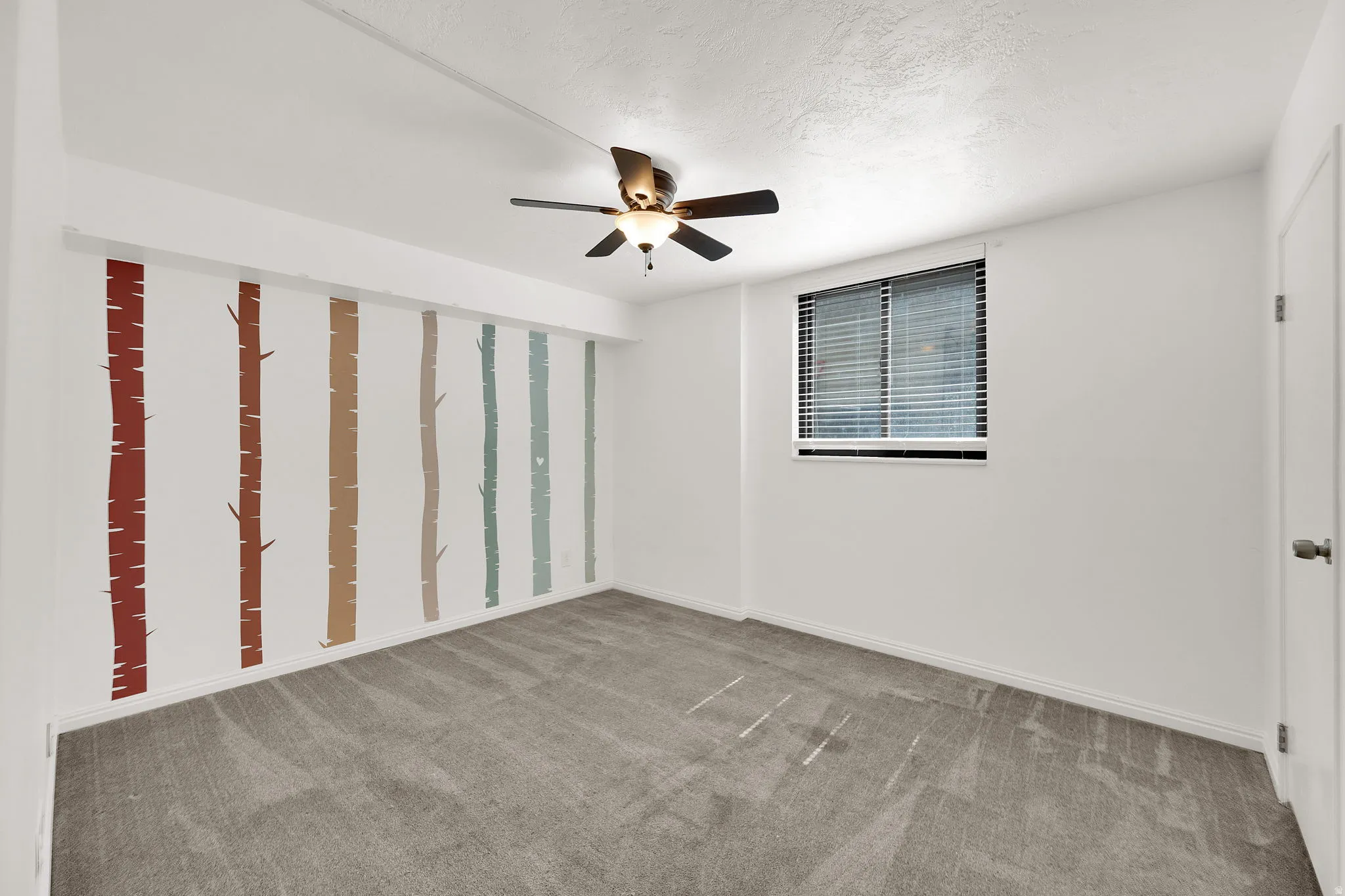 White walls with accent wall featuring colorful wall treatment, gray carpet flooring, window with blinds, and ceiling fan with integrated lighting