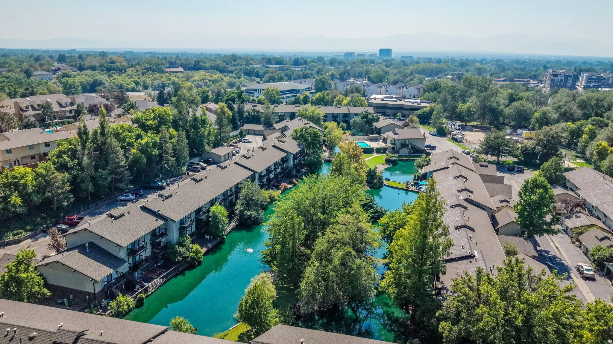 Aerial perspective showcasing multiple residential buildings, surrounded by mature trees, and featuring a prominent water feature