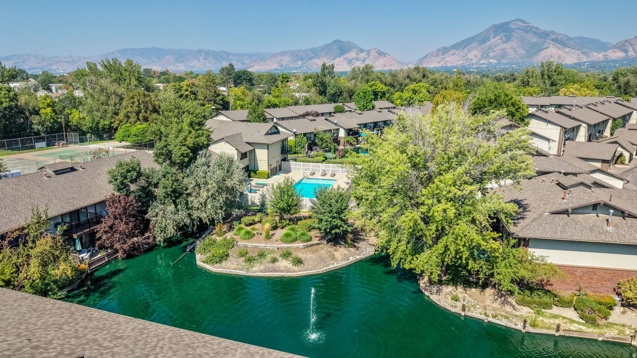 Community setting with a central pond featuring a fountain, surrounding mature trees, a swimming pool, and tennis courts
