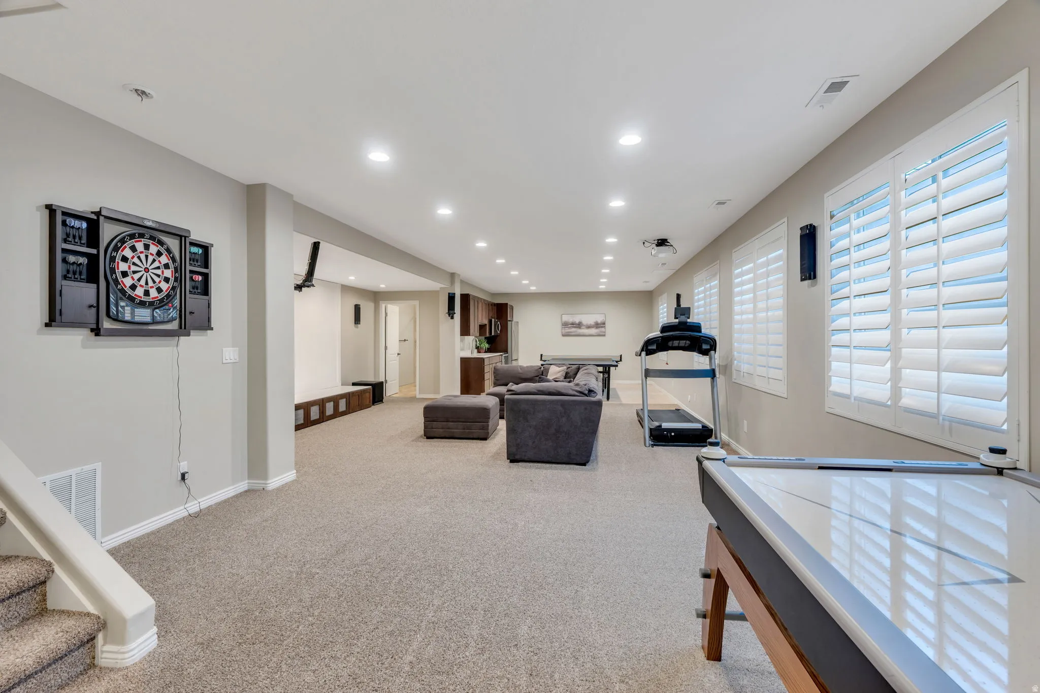 Expansive lower-level recreational space featuring recessed lighting, a carpeted floor, multiple windows with white plantation shutters, and an integrated entertainment system