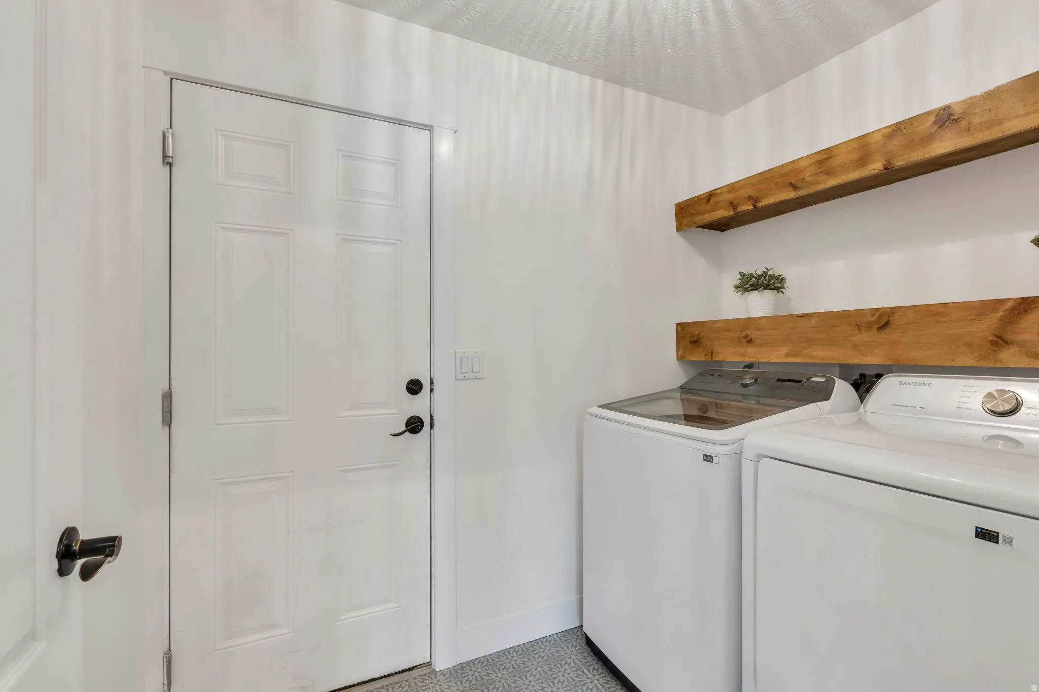 Dedicated laundry area featuring a paneled door with matte black hardware, wood-finish floating shelves, and patterned tile flooring