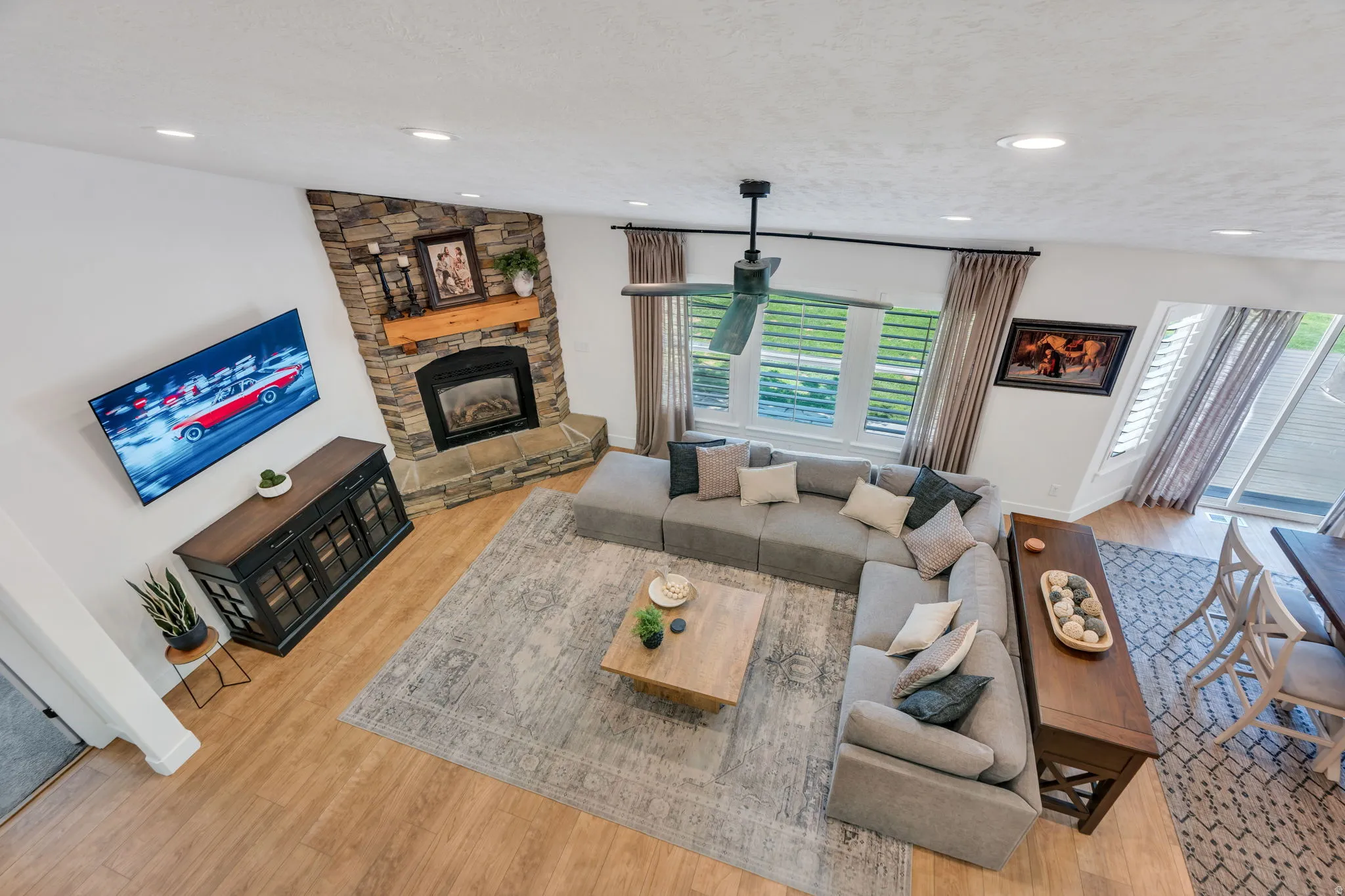 Spacious living area featuring a corner stone fireplace with a wood mantle, wood-finish flooring, recessed lighting, a ceiling fan, and large windows with horizontal blinds