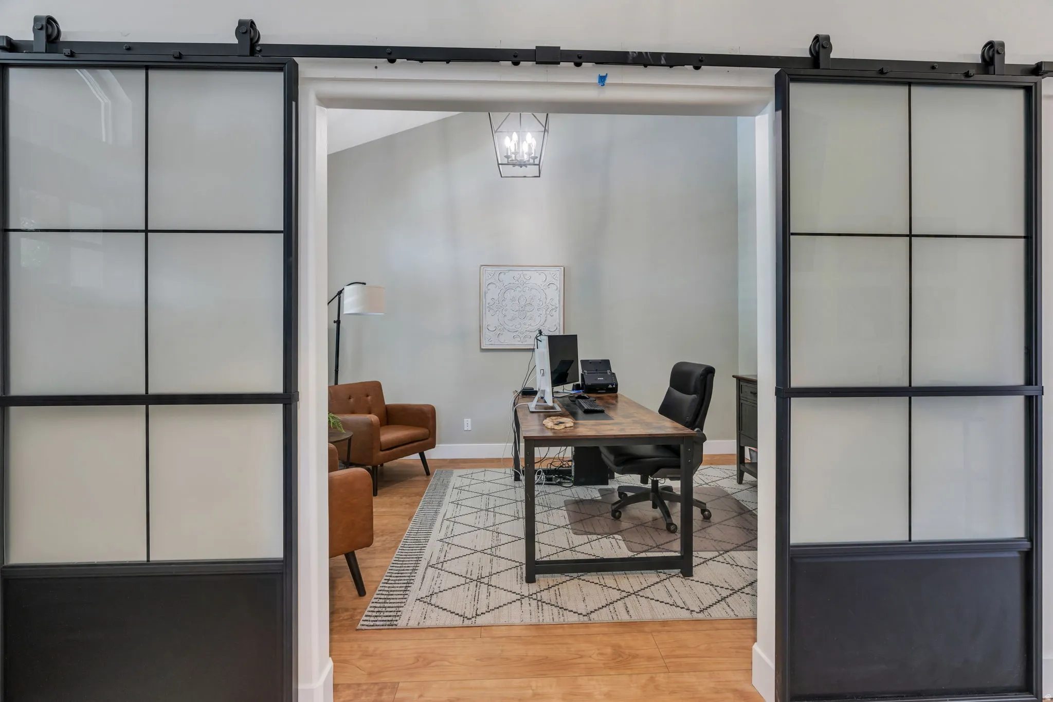 Dual black framed sliding barn doors with frosted glass panels open to a room featuring wood-finish flooring, light gray walls, and a contemporary chandelier