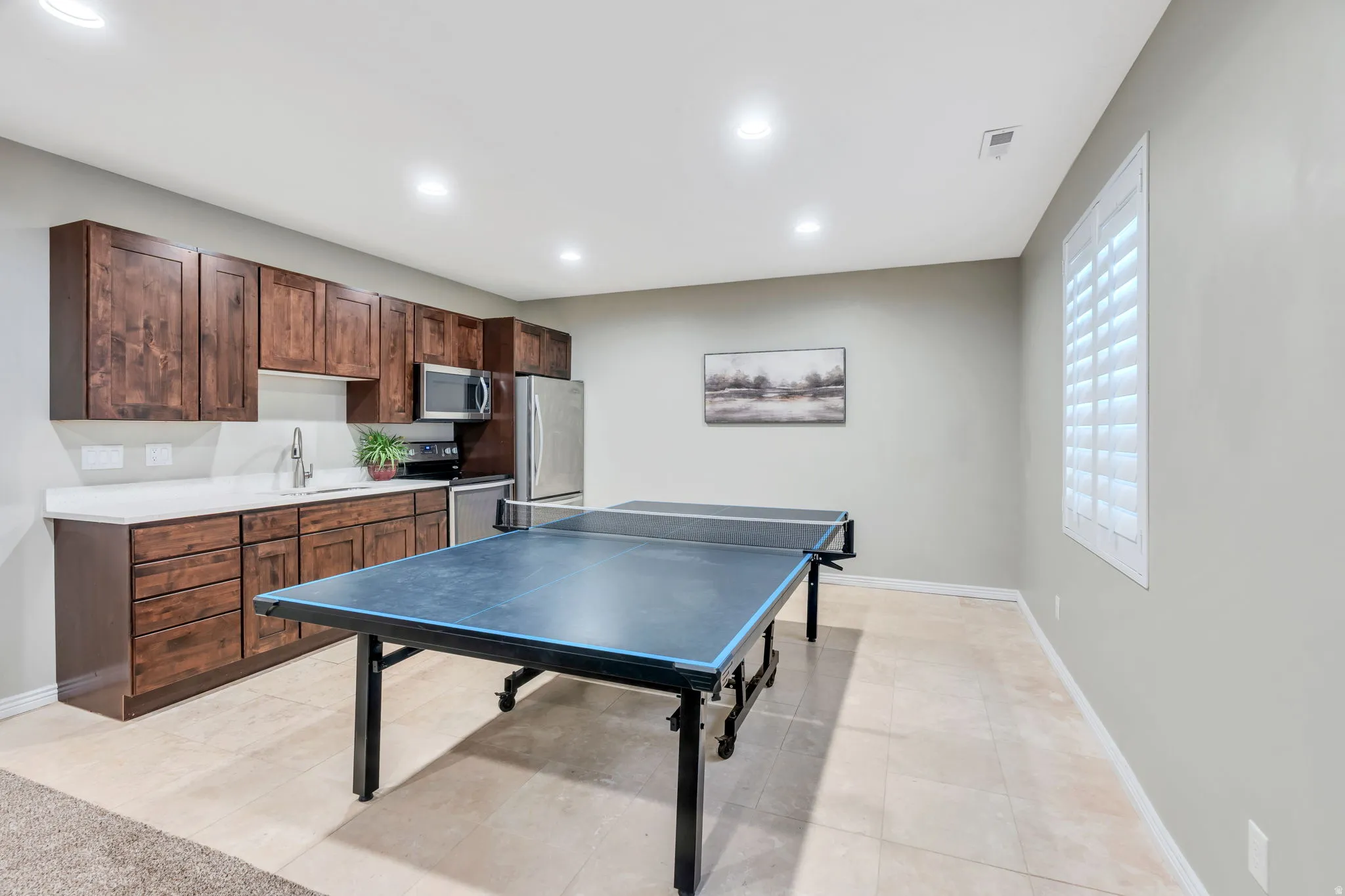 Versatile recreation room featuring a professional-grade ping pong table, wood-finish cabinetry with ample storage, stainless steel appliances, white subway tile backsplash, quartz countertop, recessed lighting, and large windows with plantation shutters