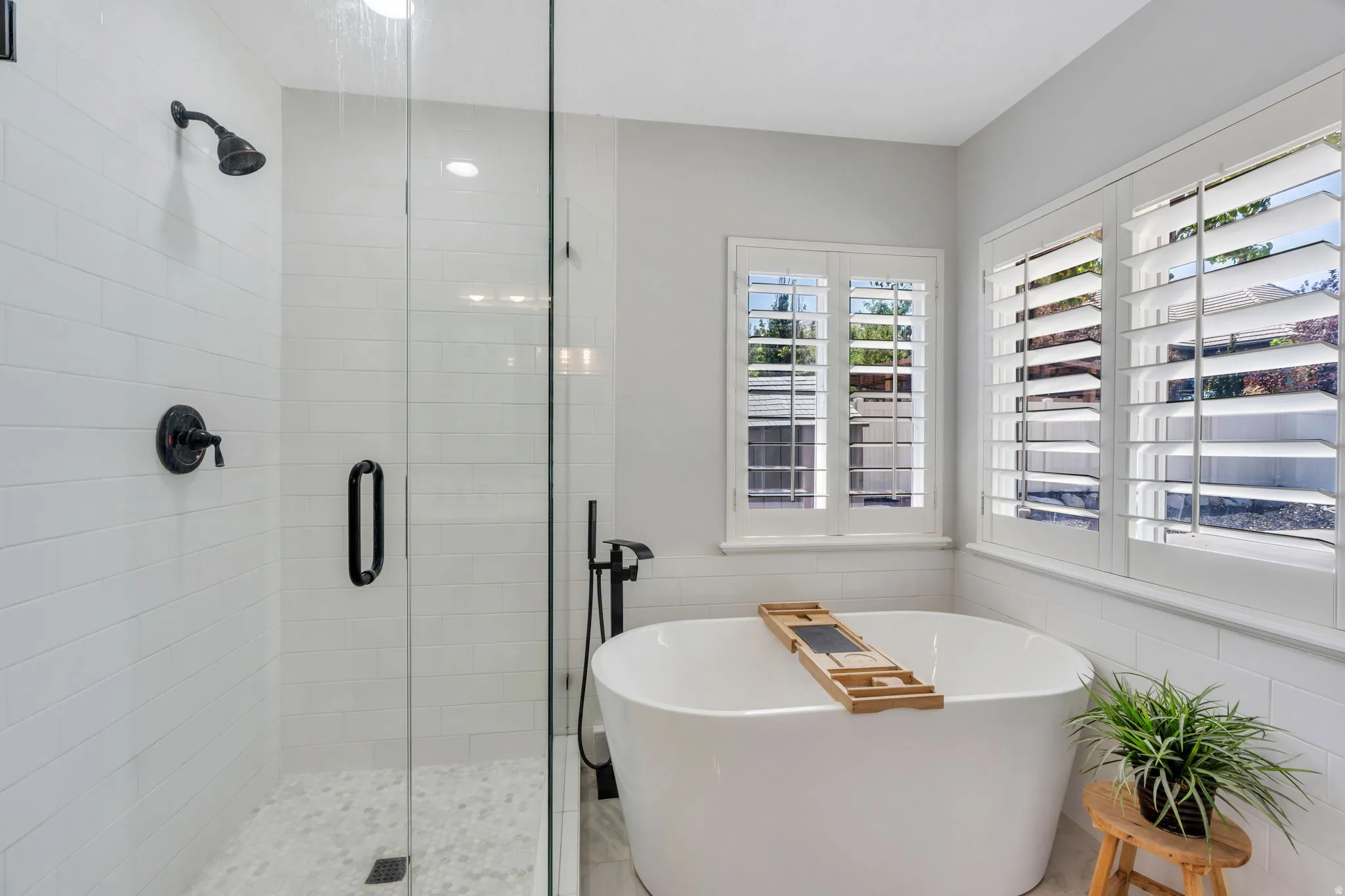 Freestanding soaking tub with plantation shutters, walk-in shower featuring a glass enclosure, white subway tile surround, and pebble-style floor