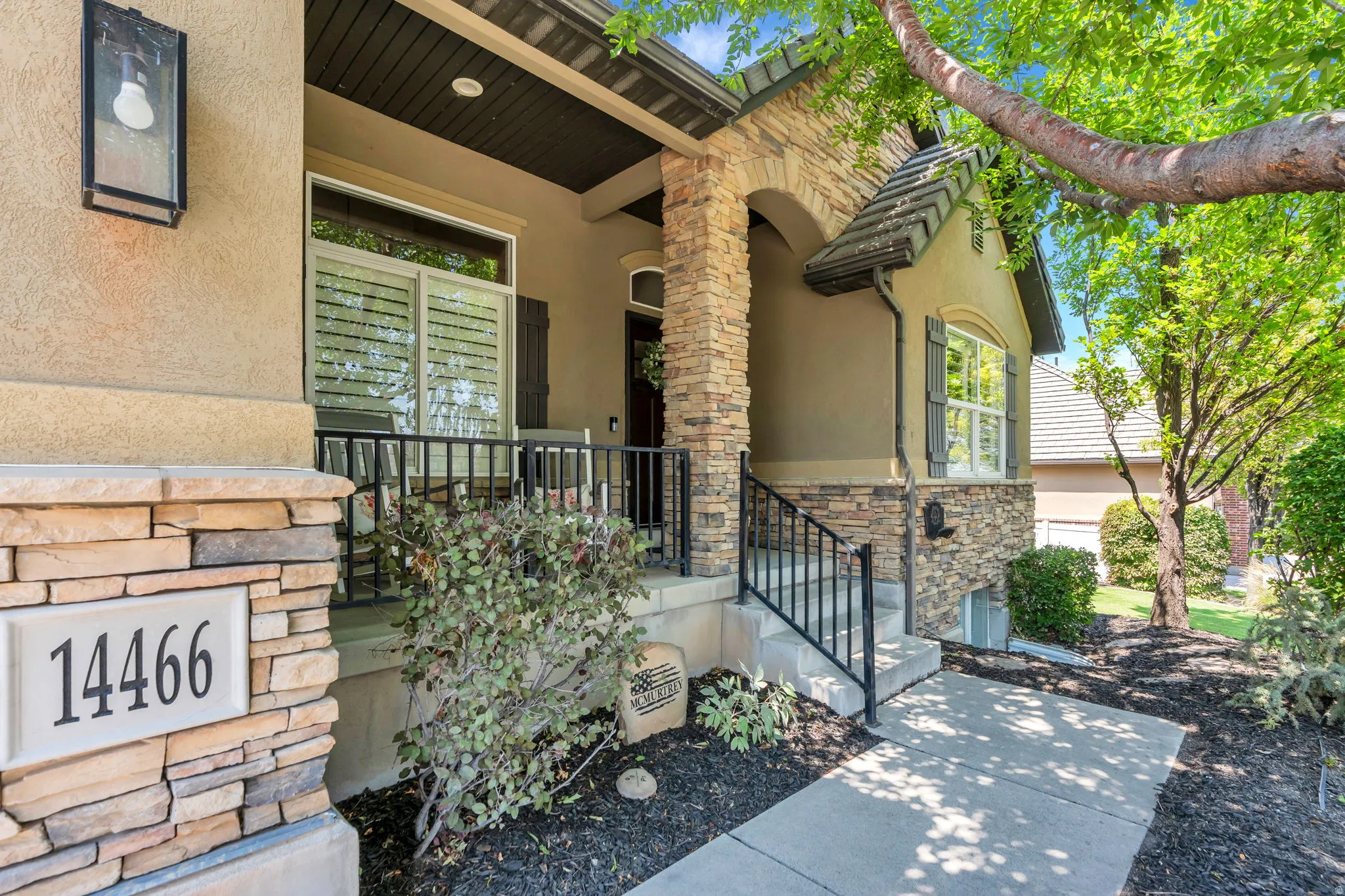 Stone and stucco exterior featuring a covered front porch with black metal railings, a concrete walkway, and established landscaping