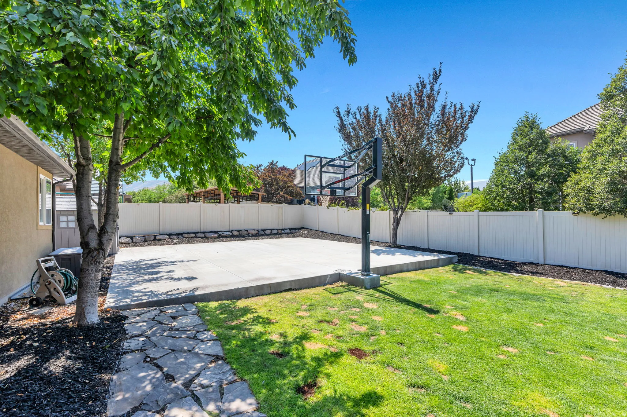 Expansive backyard featuring a concrete sport court with an attached basketball hoop, a manicured lawn, a flagstone pathway, mature trees, and a white privacy fence