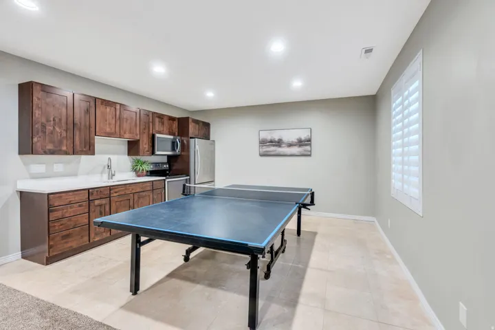 Versatile recreation room featuring a professional-grade ping pong table, wood-finish cabinetry with ample storage, stainless steel appliances, white subway tile backsplash, quartz countertop, recessed lighting, and large windows with plantation shutters