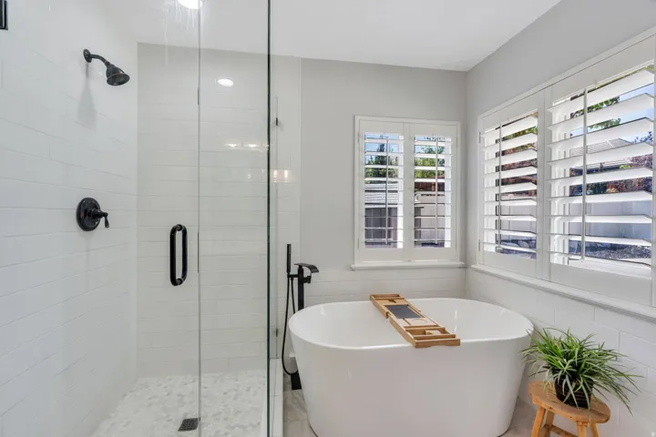 Freestanding soaking tub with plantation shutters, walk-in shower featuring a glass enclosure, white subway tile surround, and pebble-style floor