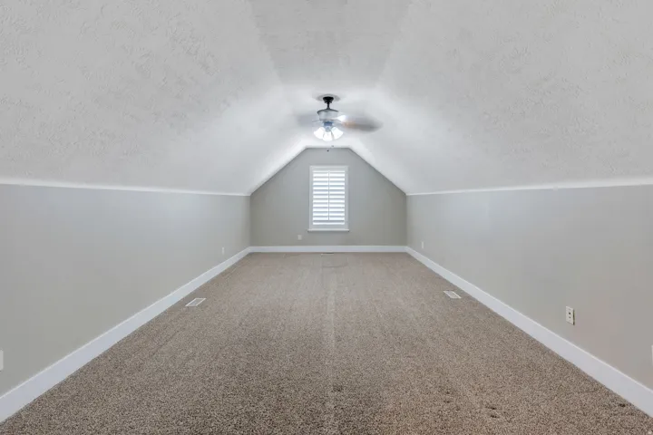 Carpeted loft space featuring sloped ceilings, a ceiling fan with light fixture, a window with louvered shutters, and baseboard trim