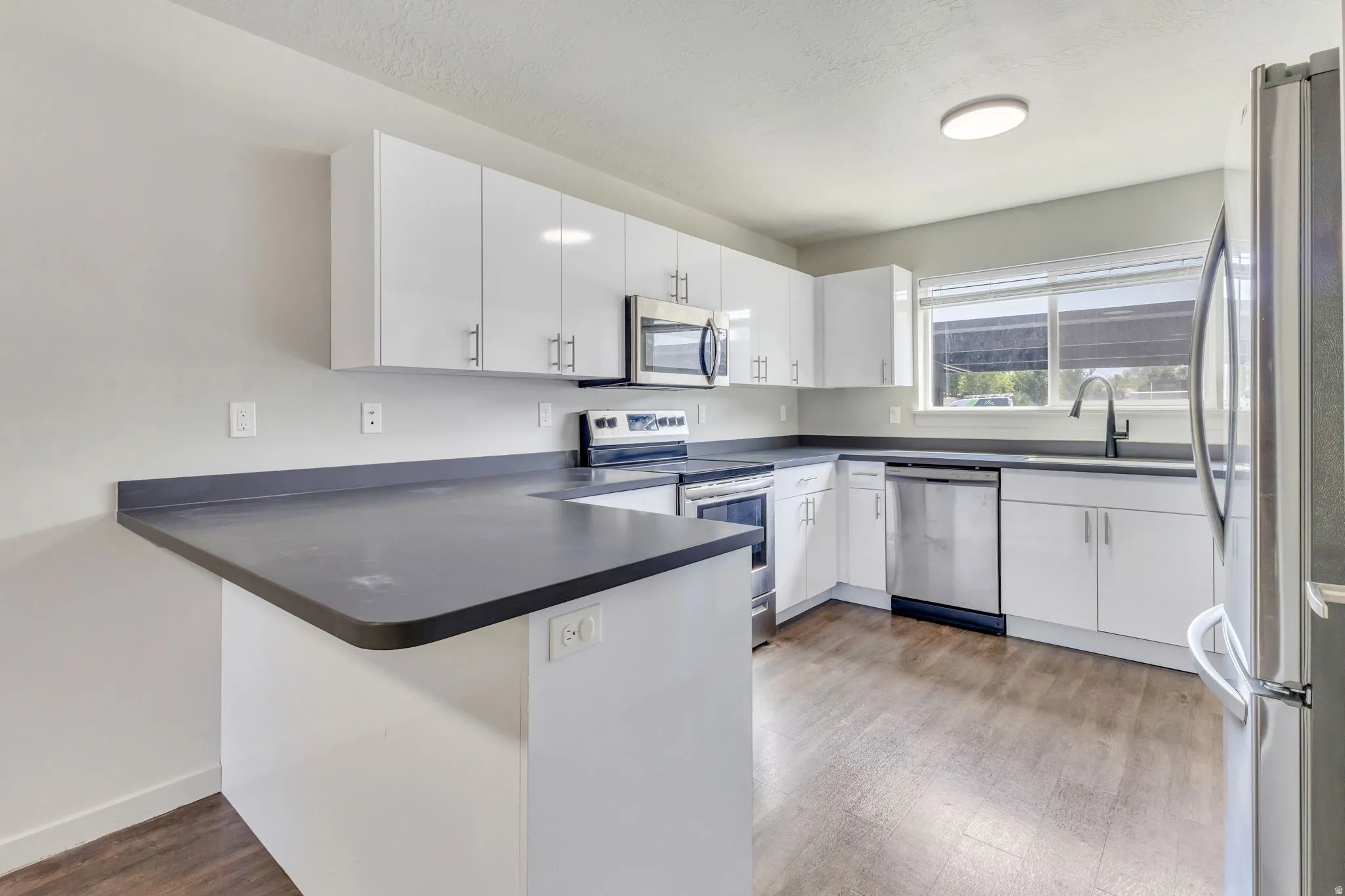 Kitchen featuring white cabinetry, dark countertops, stainless steel appliances, wood-finish flooring, and a window with blinds