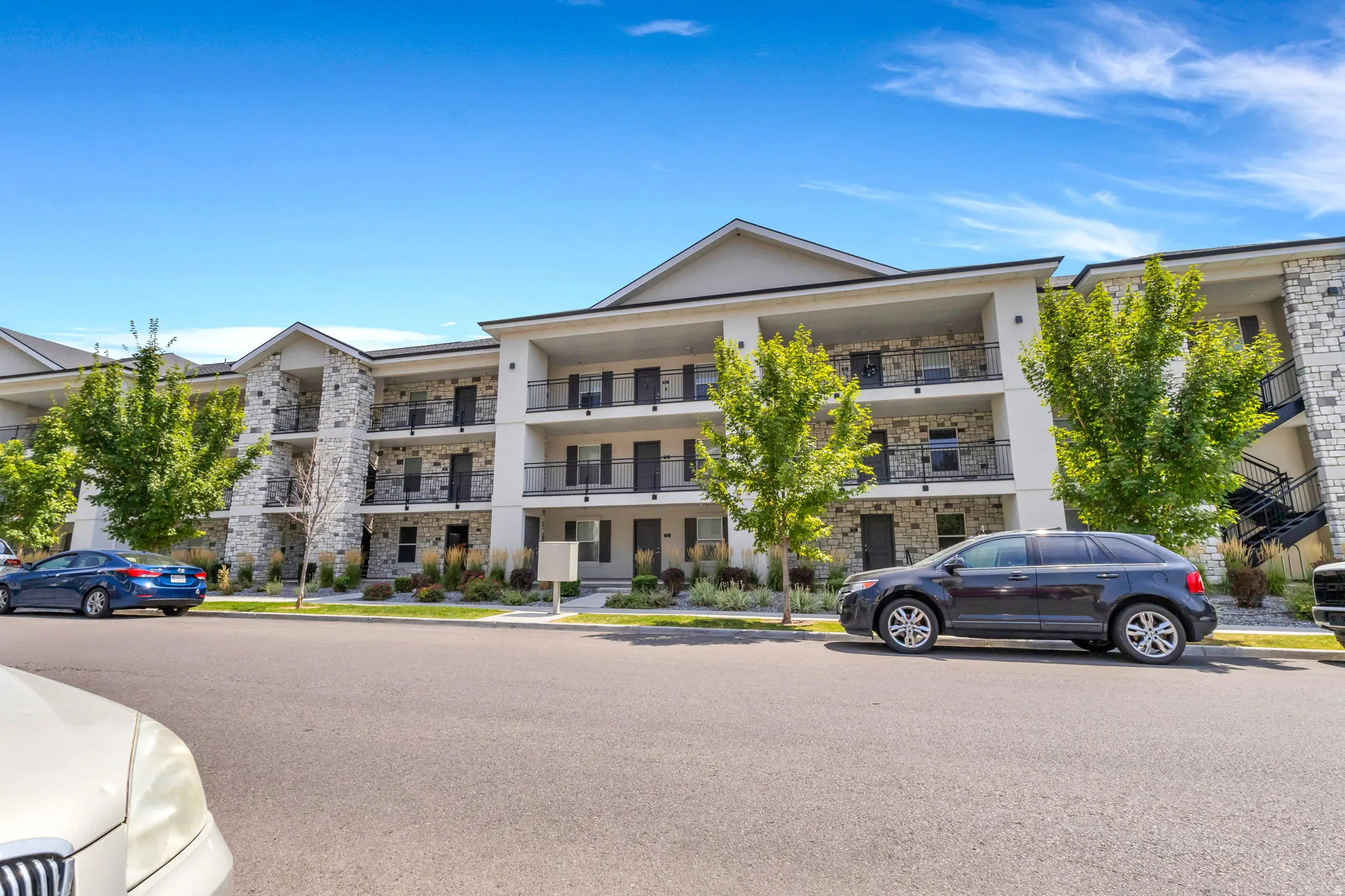 Multi-story building featuring stone veneer accents, multiple balconies with black railings, and landscaping with mature trees