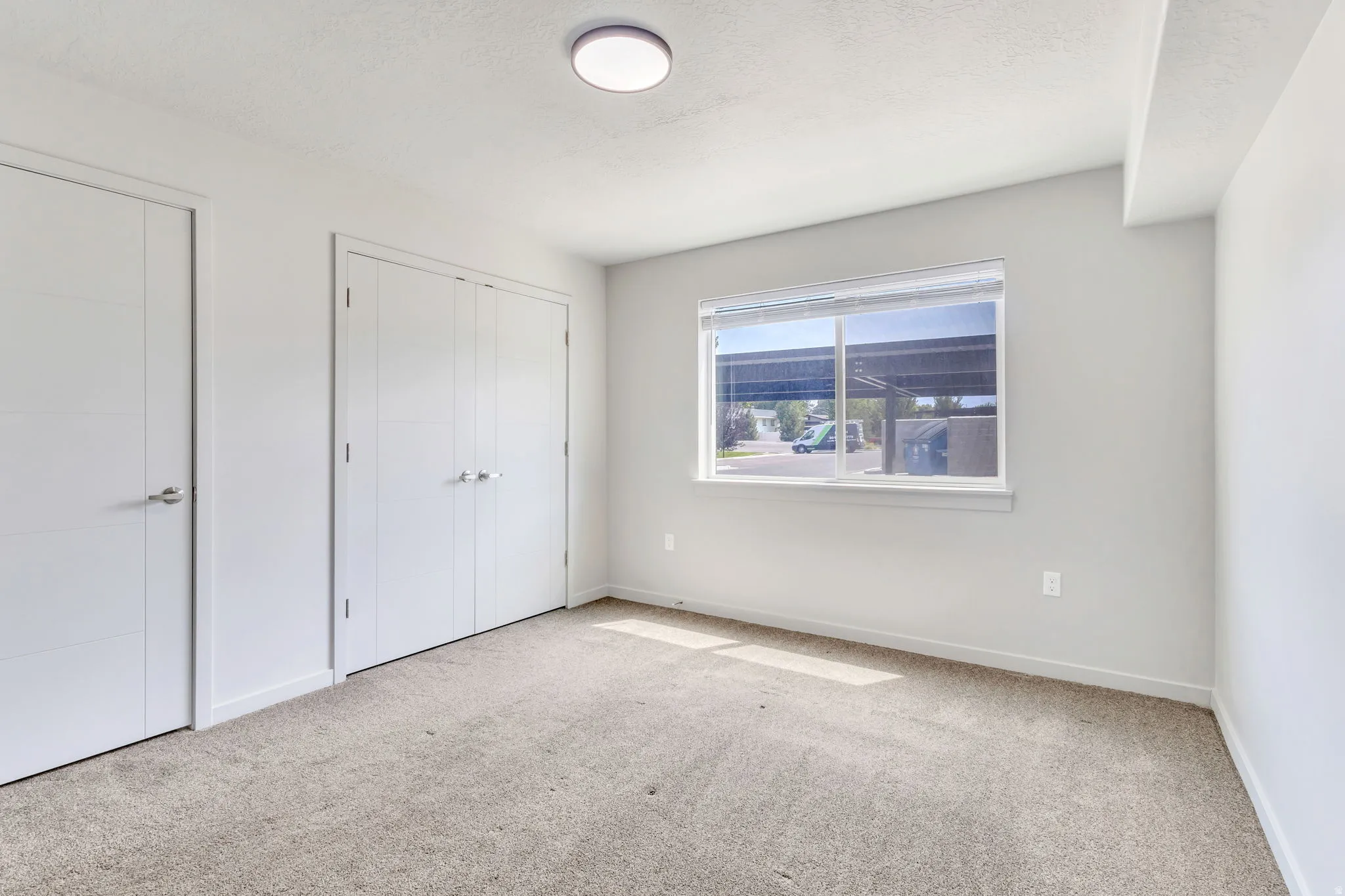 Bright, neutral bedroom featuring neutral carpet flooring, dual closets with white doors, a large window with natural light, and clean white walls with recessed ceiling lighting