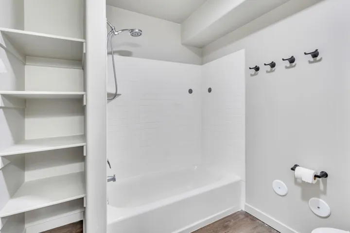 Bathtub with white subway tile surround and shower fixture, built-in shelving, dark-finish wall hooks, and wood-finish flooring