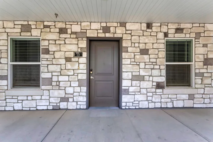 Stone veneer exterior with a paneled entry door, two windows with blinds, and a concrete floor