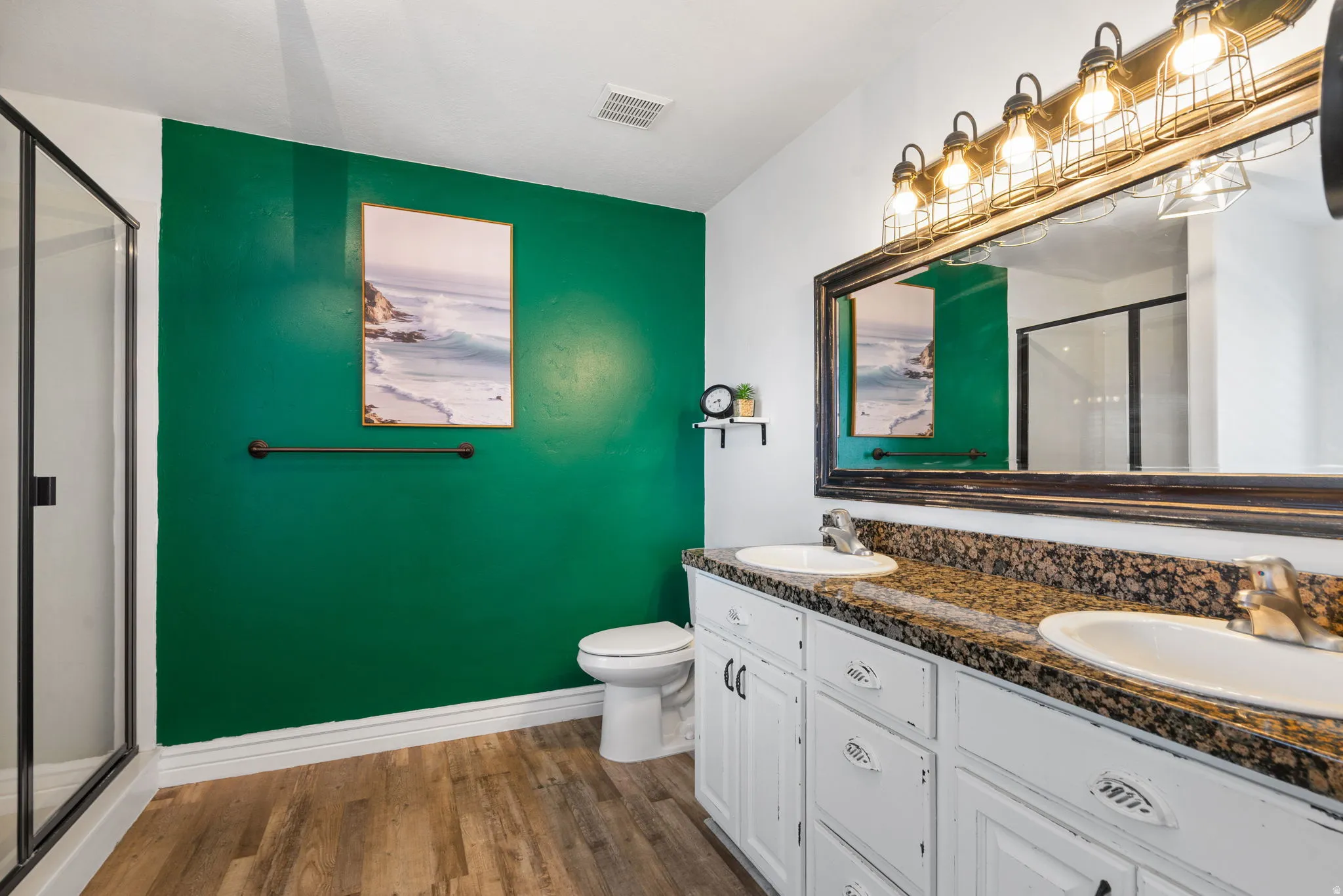 Dual vanity with integrated sinks, wood-finish flooring, glass-enclosed shower, white cabinetry, and granite-style countertop