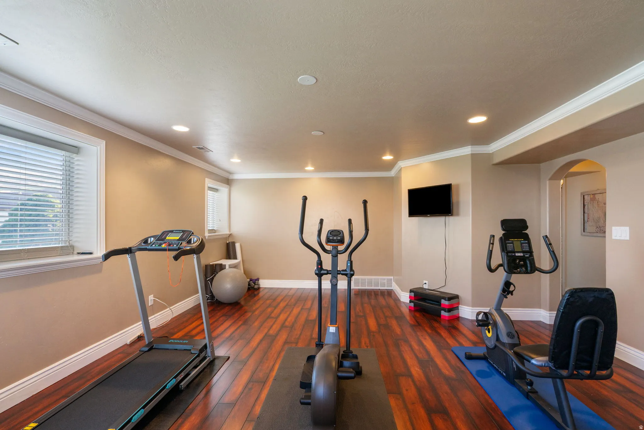 Dedicated home fitness room featuring recessed ceiling lighting, wood-finish flooring with blue accent runner, mounted flat-screen television, and multiple exercise stations including treadmill, stationary bike, and elliptical equipment