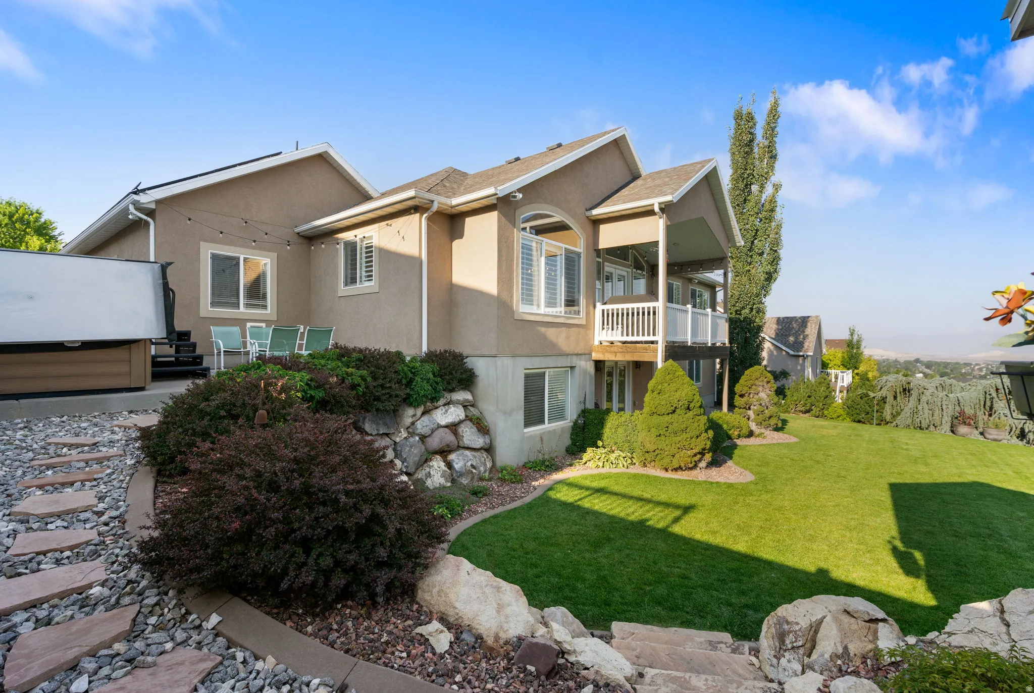 Multi-level stucco exterior with an upper-level covered balcony, lower-level patio, and a terraced backyard featuring a manicured lawn