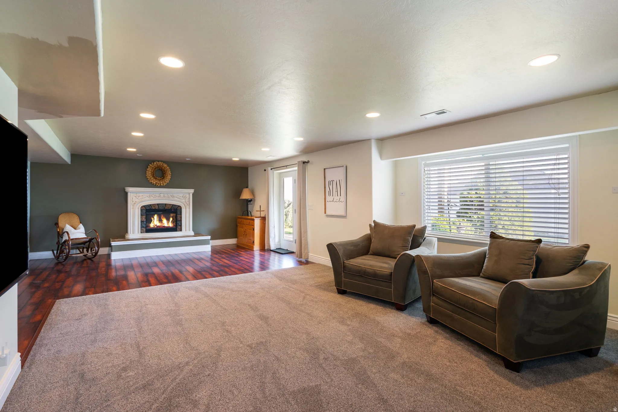 Recessed lighting illuminates a spacious room featuring a decorative fireplace with a carved mantel, wood-finish flooring, and a large window with blinds