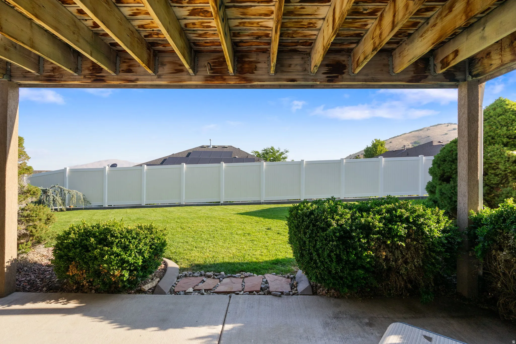 Covered patio with concrete flooring, overlooking a green lawn with established shrubs and a white privacy fence