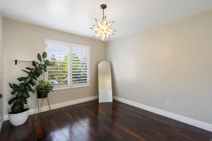 Bright and spacious room showcasing dark wood-finish flooring, large windows with plantation shutters offering natural light and privacy, and a decorative brass star pendant fixture