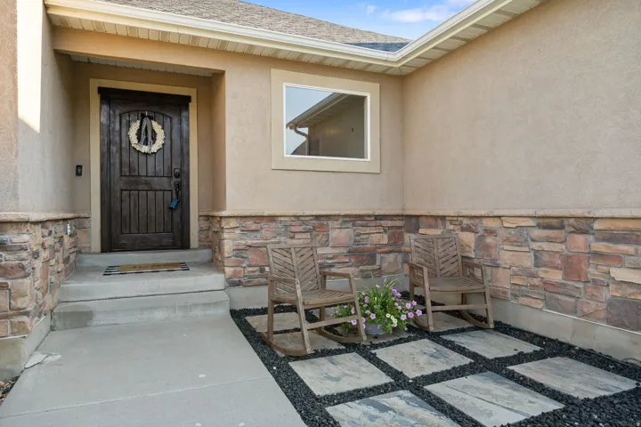 Welcoming entry portico featuring a decorative wreath-adorned wood plank door with sidelite window, mixed-material exterior combining stucco upper walls with stacked stone veneer wainscoting, geometric hardscape patterning with contrasting stone and dark