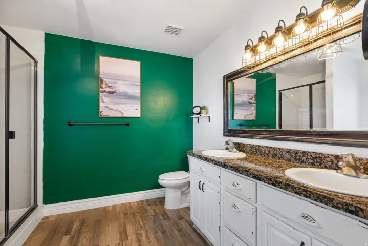 Dual vanity with integrated sinks, wood-finish flooring, glass-enclosed shower, white cabinetry, and granite-style countertop