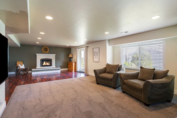 Recessed lighting illuminates a spacious room featuring a decorative fireplace with a carved mantel, wood-finish flooring, and a large window with blinds