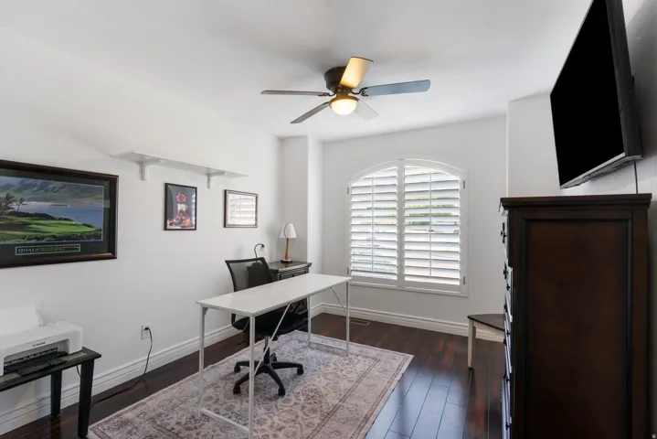 Wood-finish flooring, white walls, arched window with plantation shutters, ceiling fan with integrated lighting, and built-in wall shelving