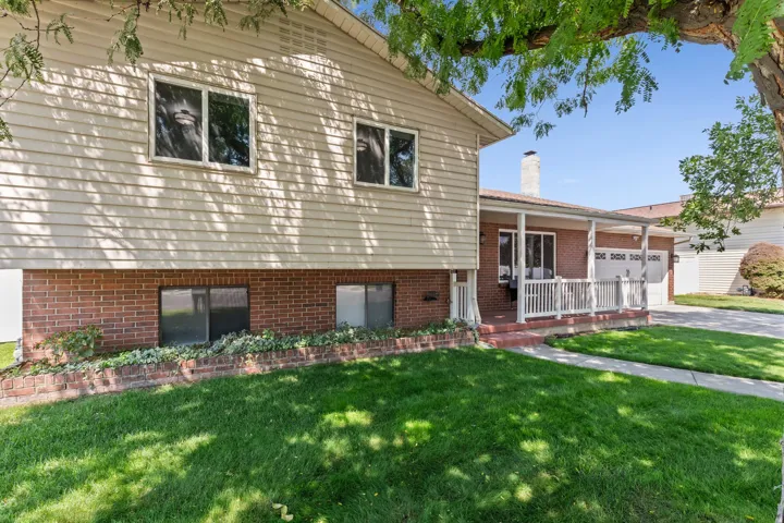 Split-level home featuring a manicured front lawn with mature shade trees, brick foundation with horizontal siding upper level, multi-pane windows, and an attached deck with white railings visible to the side