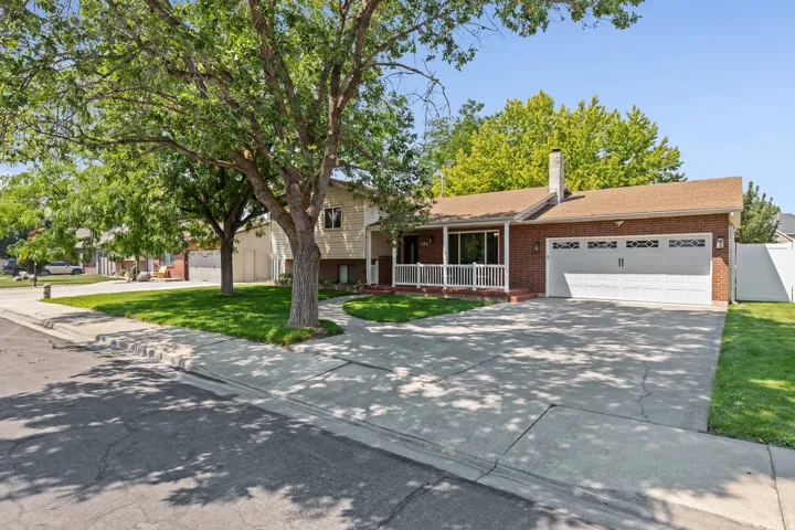 Brick colonial with white-trimmed front porch, mature shade tree anchoring the landscaped front lawn, two-car garage with white panel doors, and concrete driveway