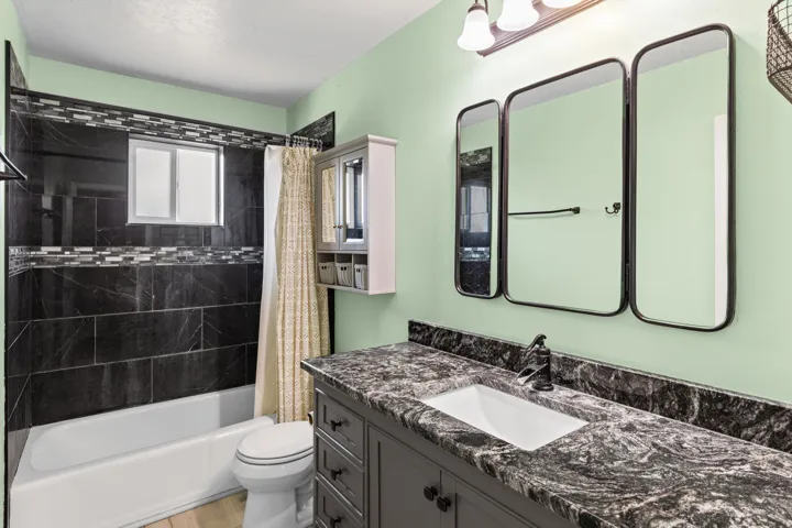 Vanity with a patterned countertop and integrated sink, dark wood-finish cabinetry, three individual framed mirrors, and a shower with dark tiled walls and a decorative mosaic border