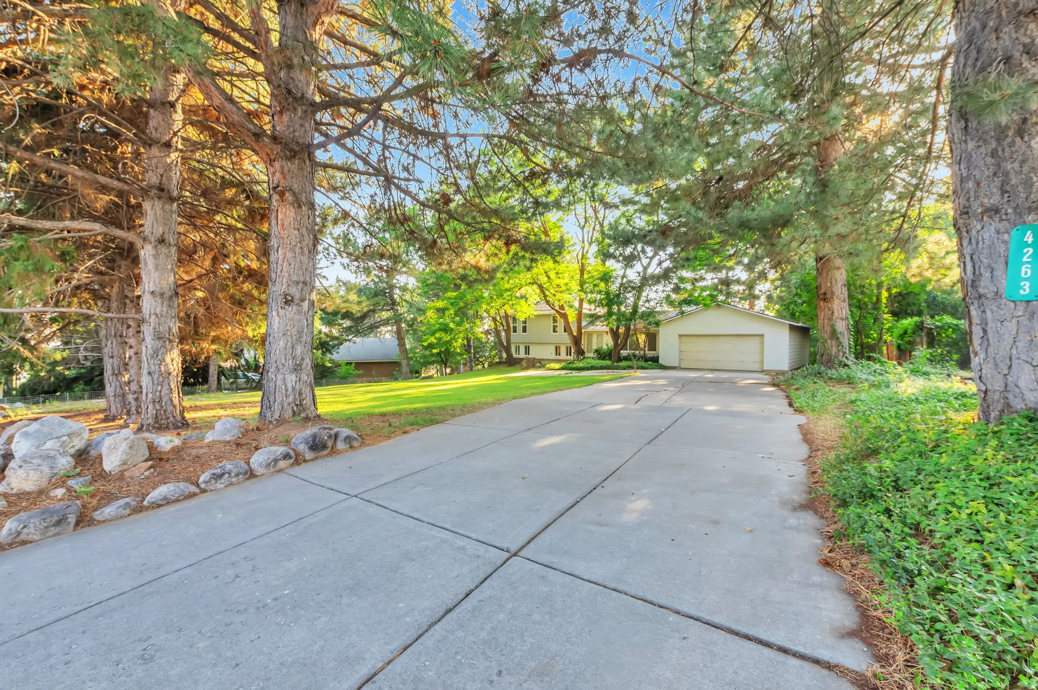 Concrete driveway with a detached two-car garage, mature trees, and natural stone landscaping elements