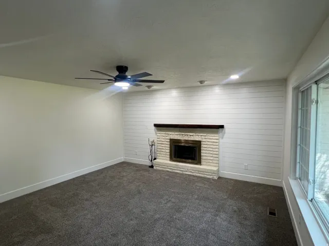 Carpeted living space featuring a fireplace with a stone surround, a wood mantle, shiplap accent wall, and a ceiling fan with integrated lighting