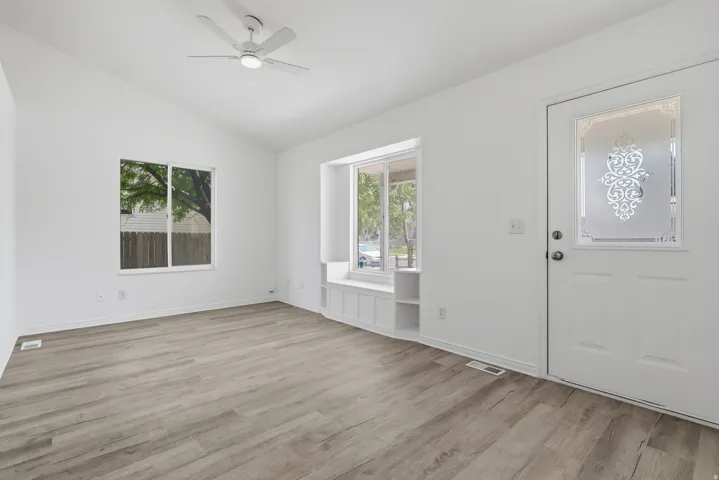 Spacious room featuring vaulted ceilings, wood-finish flooring, a built-in window seat with storage, a decorative glass-paneled entry door, and a ceiling fan
