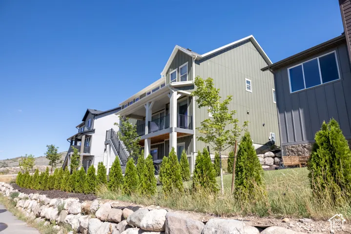 Multi-level exterior featuring vertical siding, a covered balcony with metal railing, stone retaining walls, and landscaped garden beds with evergreen shrubs