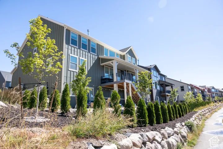 Multi-story home exterior featuring vertical panel siding, elevated balcony with black metal railing, stone retaining wall, and landscaped perimeter with young trees