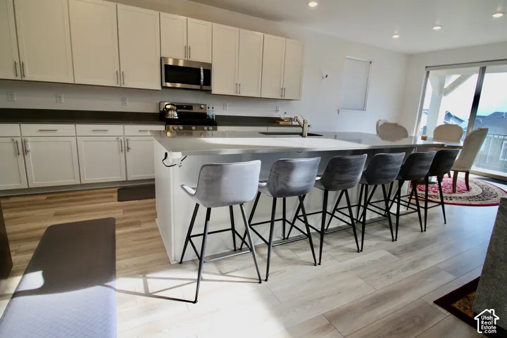 Open-concept kitchen featuring an expansive central island with seating, white shaker-style cabinetry, stainless steel appliances, and light wood-finish flooring