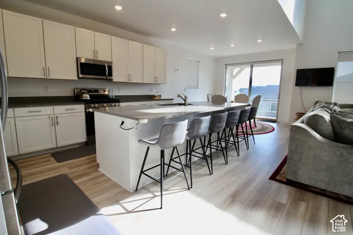 Open-concept kitchen and living area featuring a large center island with seating for six, light-toned wood-finish flooring, white cabinetry, dark countertops, and a sliding glass door to exterior