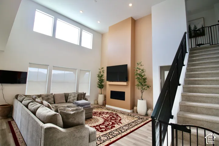 Open-concept living space featuring high ceilings, recessed lighting, wood-finish flooring, a contemporary fireplace, and a carpeted staircase with dark metal railings