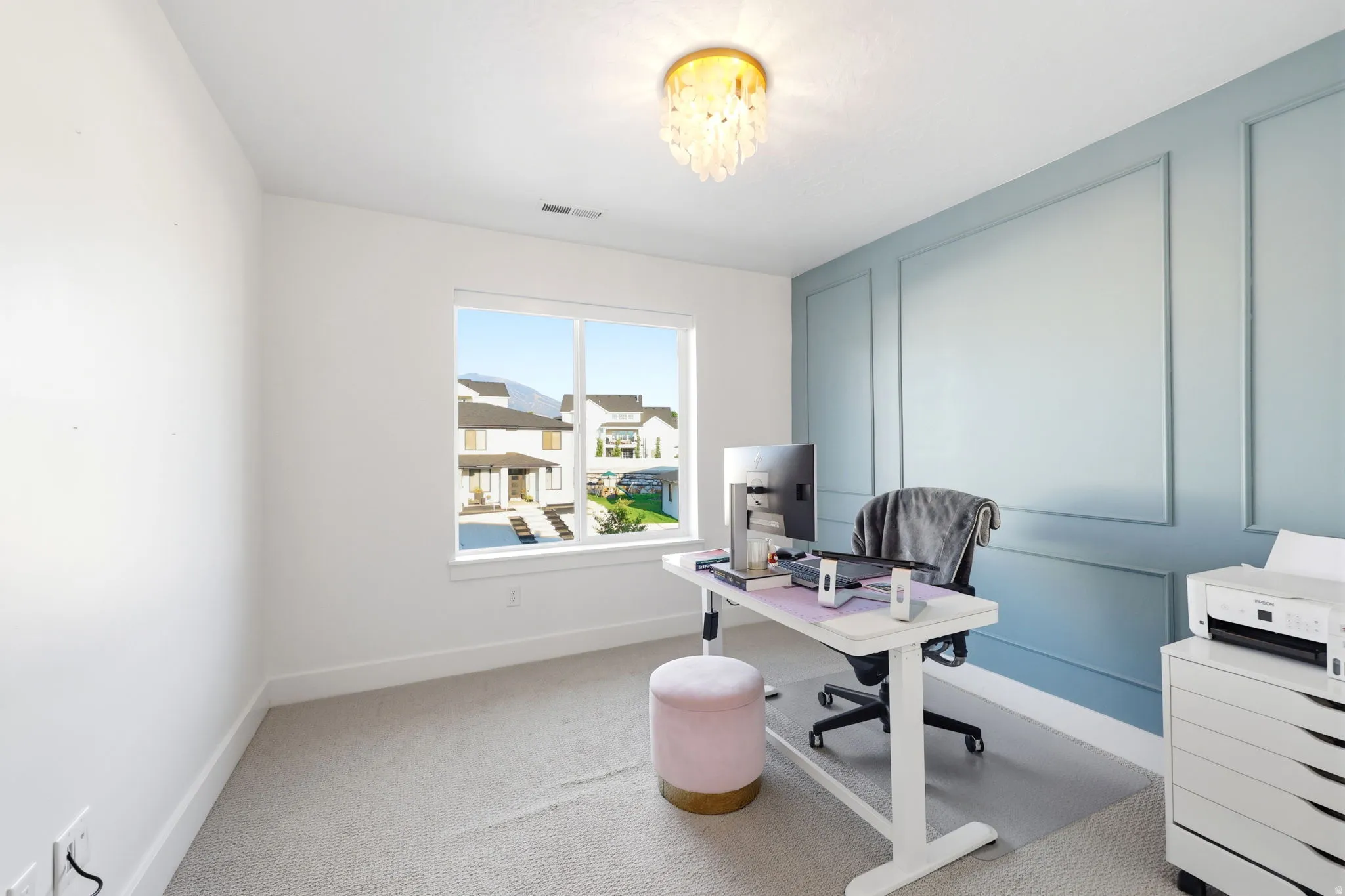 Bright, naturally lit home office featuring expansive windows with neighborhood sightlines, light wood-finish flooring, and soft gray wall paneling