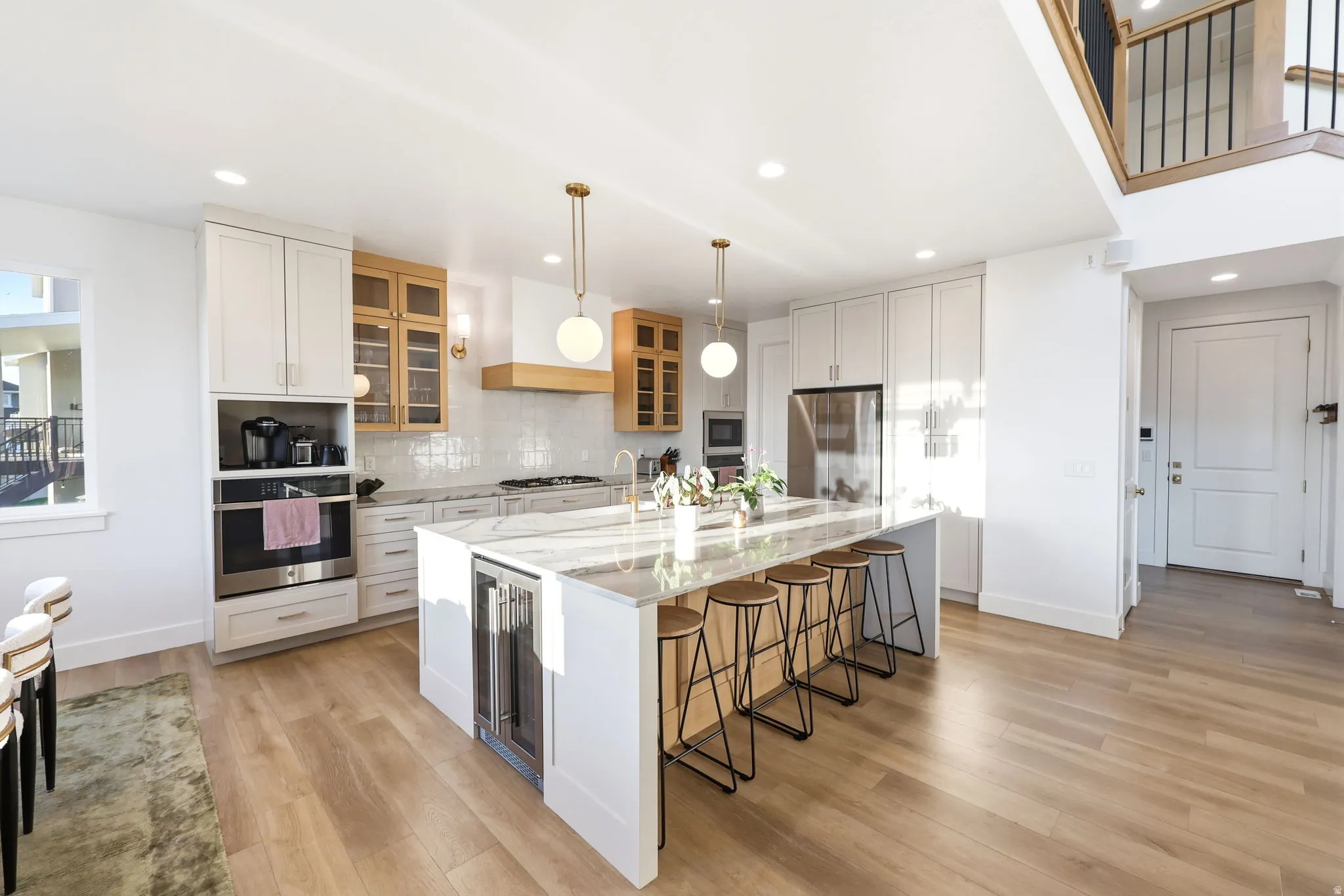 Modern kitchen featuring a large island with integrated beverage cooler, light wood-finish flooring, white upper and lower cabinetry, stainless steel appliances, and a tile backsplash