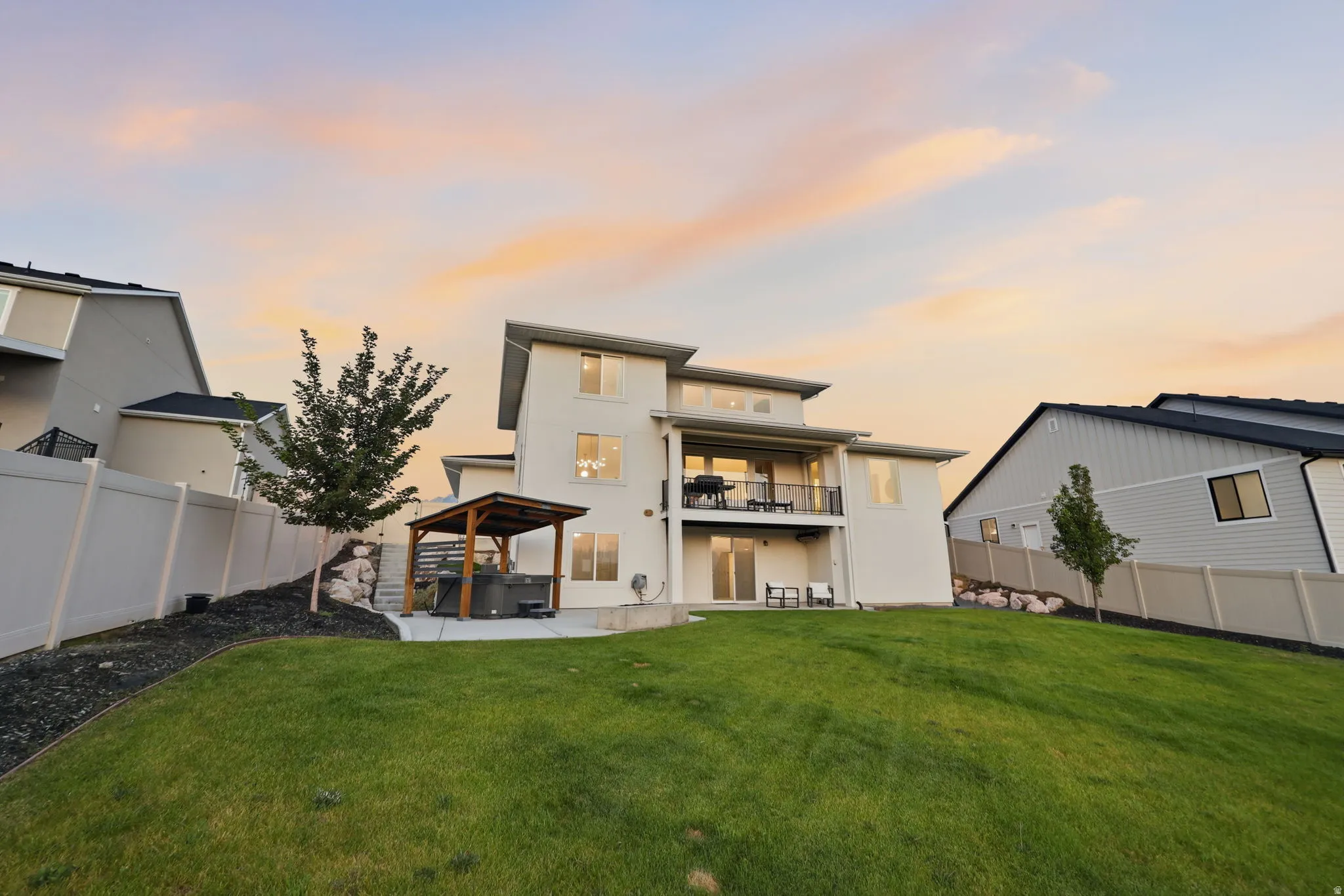 Modern two-story residence showcasing a spacious rear elevation with expansive manicured lawn, contemporary architectural lines, multi-level deck with integrated outdoor living space, large windows with evening illumination, and professionally landscaped