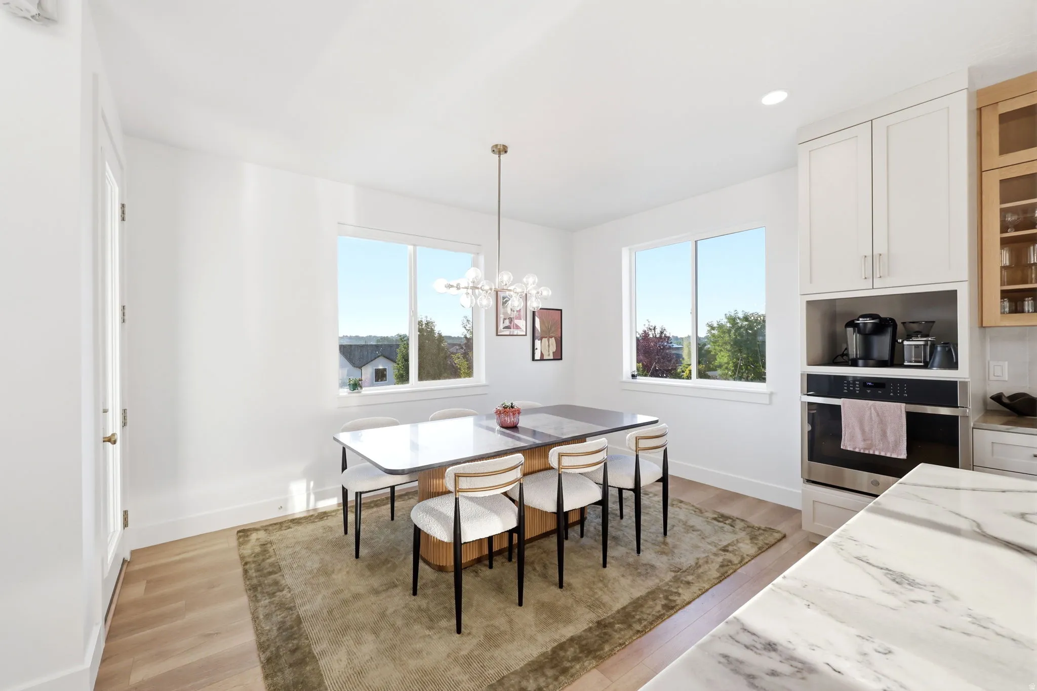 Bright, light-filled dining area featuring a wood-finish dining table with coordinating seating, positioned beneath dual large windows framing natural landscape views